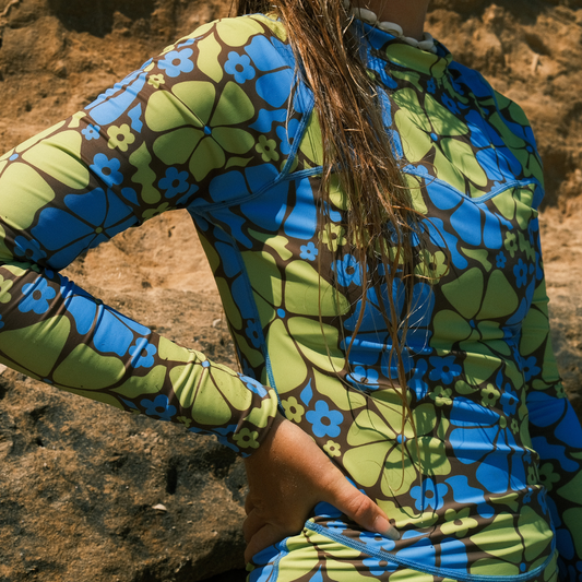 A person wearing a multicolored surf suit with a floral pattern, sitting on a rock by the beach.
